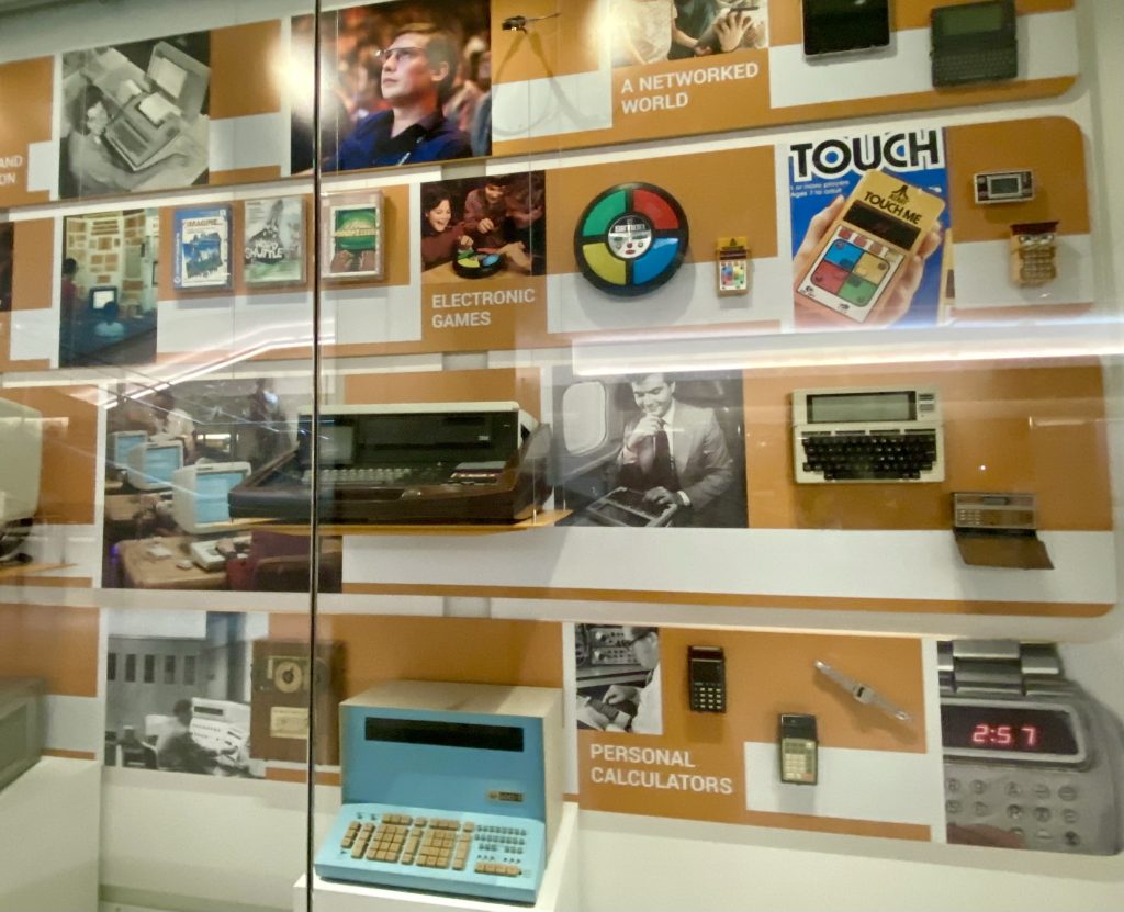 Display case featuring vintage electronics, including calculators, typewriters, electronic games, and related historical items, arranged by category against orange background panels.