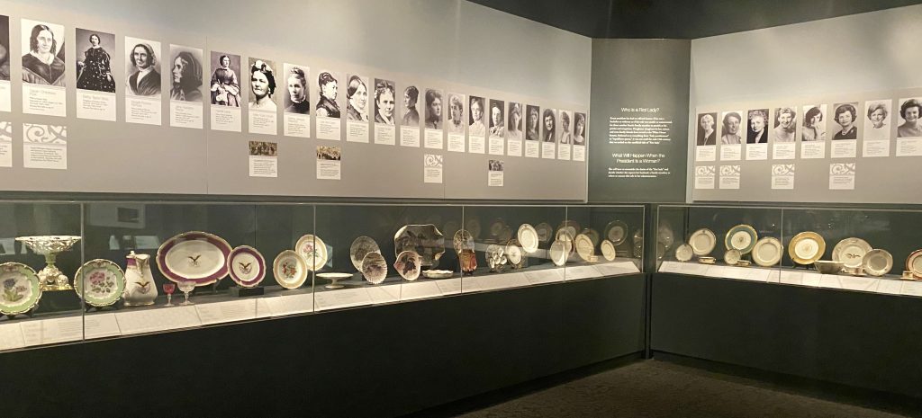 Numerous, unique dinner place settings in glass display case with pictures of first ladies above