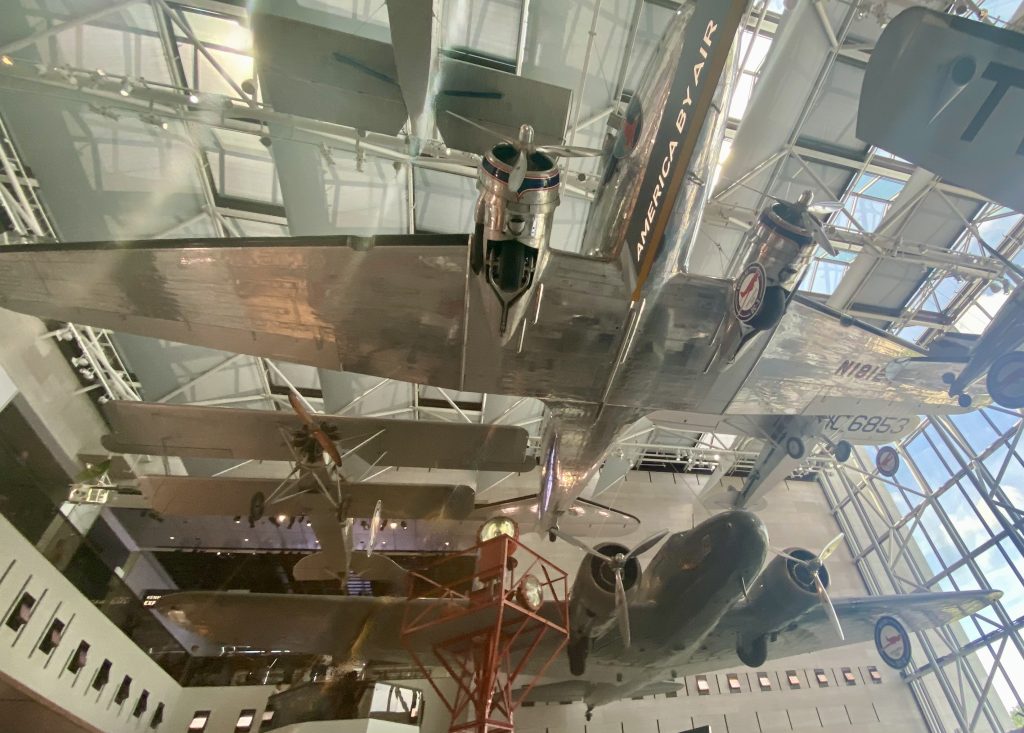 Several planes hanging from the ceiling of the National Air and Space Museum in Washington DC - a small biplane, a large military aircraft, a large silver plane that says "America by Air"