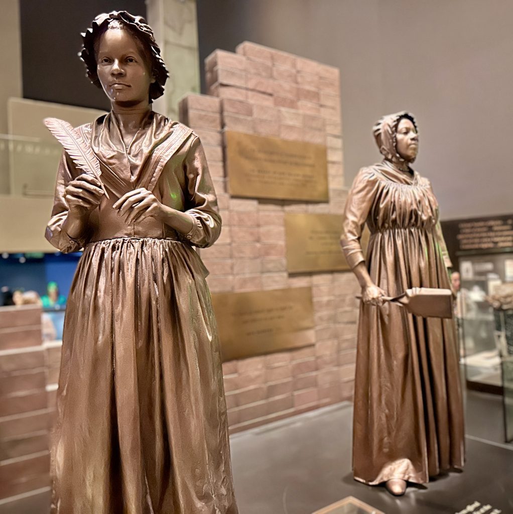 two bronze statues of African American women, one holding a feather pen and one holding a dust pan