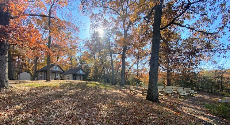Two small cabins sitting on top of a hill among trees with orange and red leaves, several sitting benches in foreground